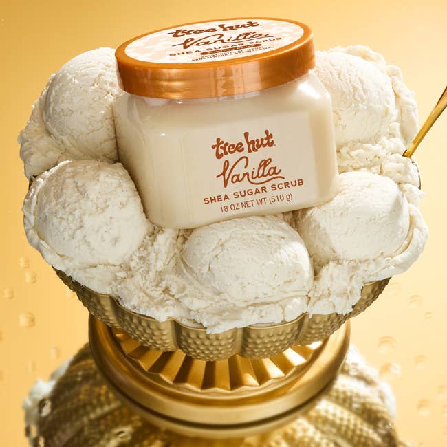 Tree Hut Vanilla Shea Sugar Scrub jar connected  a golden  stand, surrounded by scoops of crystal  cream