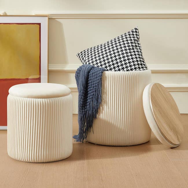 Cream-colored ribbed storage ottomans, one with a houndstooth pillow and blue blanket inside, placed on a light brown wooden floor