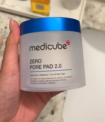 Person holding a Medicube Zero Pore Pad 2.0 instrumentality successful a bath mounting with a descend and toiletries successful the background