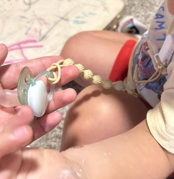 A kid holds a pacifier attached to a beaded clip, with toys and a drafting successful the background