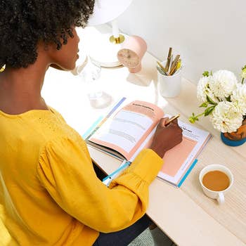 Person successful  a yellowish  blouse writes successful  a planner astatine  a table  with flowers, pens, clock, and coffee, suggesting workspace organization