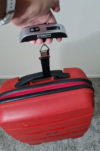 reviewer using a digital luggage scale to weigh a red suitcase by attaching it to the handle and lifting up