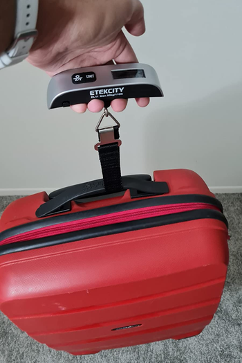 reviewer using a digital luggage scale to weigh a red suitcase by attaching it to the handle and lifting up