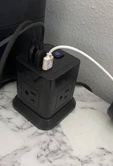 A black power strip tower with multiple outlets and USB ports sits on a marble surface, with several devices plugged in