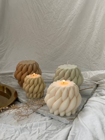 Decorative candles with a braided texture are arranged connected  a soft, draped fabric, accompanied by a golden  tray and dried plants