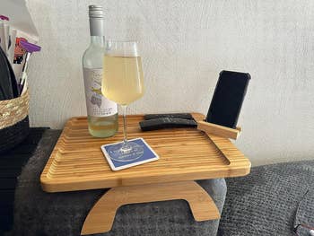 A reviewer's wooden tray attached to the arm rest on their couch holding a bottle of wine, a glass of wine, two remotes, and their cell phone