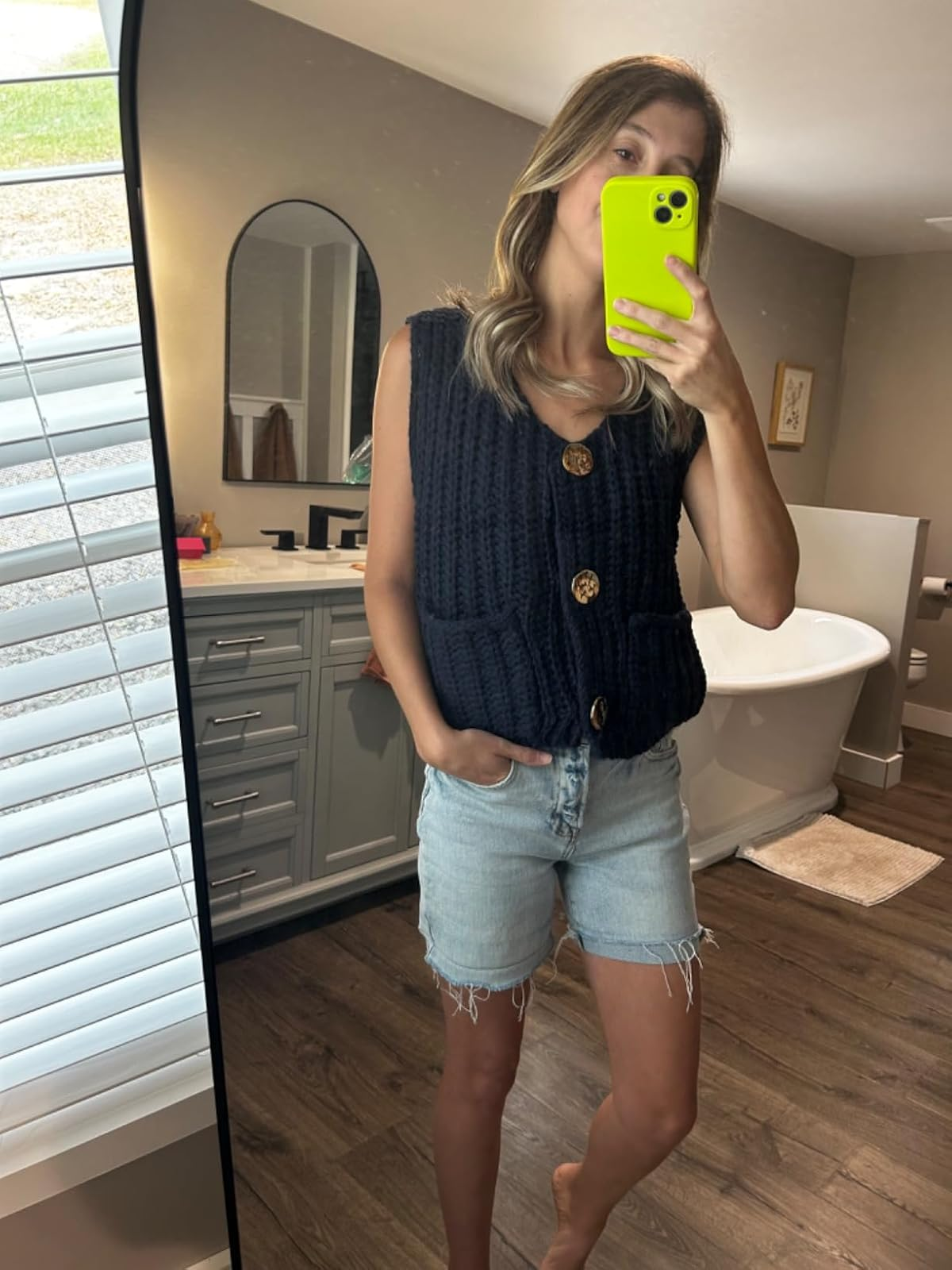 Orang yang mengambil selfie cermin mengenakan bagian atas lengan, kancing rajut dan celana pendek denim yang berjumbai, memegang telepon yang terang. Pengaturan kamar mandi di latar belakang
