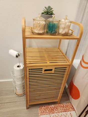 Bamboo bath  support  with a stack of toilet paper, a potted plant, and jars of fabric  balls and swabs, adjacent  to a ablution  curtain