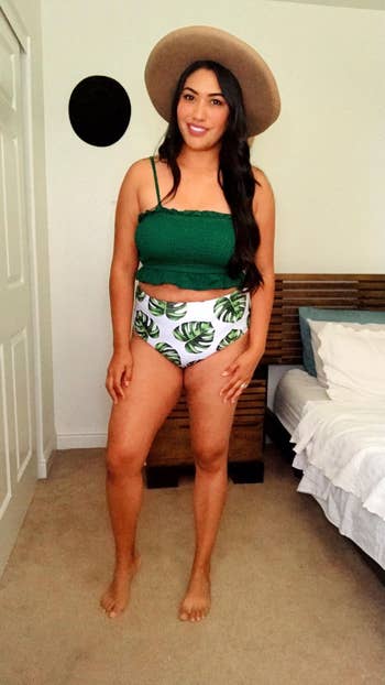 Person wearing a green crop top, leaf-patterned shorts, and a wide-brimmed hat, standing in a bedroom setting