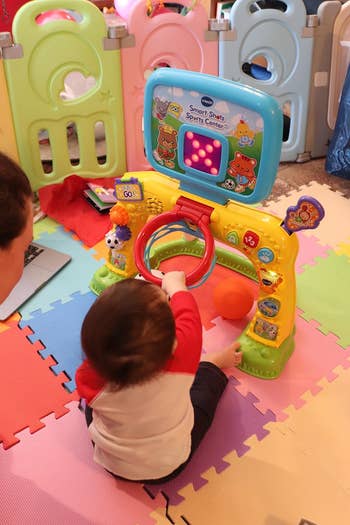A kid plays with an interactive artifact featuring buttons, lights, and sports activities, seated connected a colorful play mat