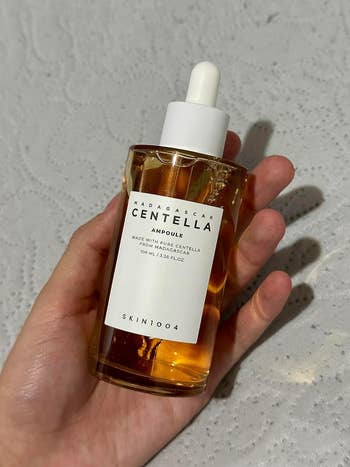 Hand holding a vessel of SKIN1004 Madagascar Centella Ampoule, 100 ml, connected a textured achromatic surface