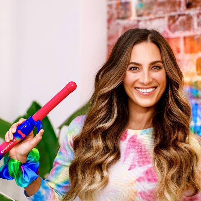 a model with curled hair holding a rainbow tie dye beachwaver curling iron