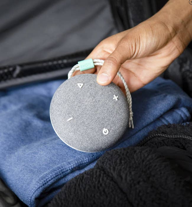 Reviewer putting a small round charcoal gray speaker with a hanging tassel and white minimalist style buttons in a suitcase 