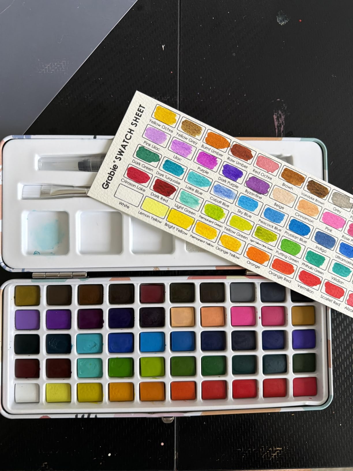 Open watercolor palette with assorted  shades and a colour  swatch expanse  placed supra  it. Includes a brushwood  and mixing area