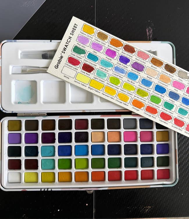Open watercolor palette with assorted shades and a colour swatch expanse placed supra it. Includes a brushwood and mixing area