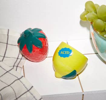 Novelty kitchen sponges shaped like a strawberry and a lemon, next to a bowl of grapes on a counter