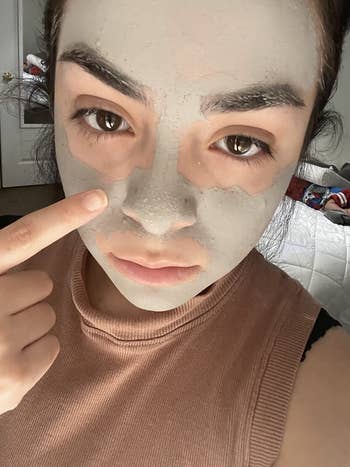 Person applying a clay face mask, pointing to their cheek, indoors for skincare shopping article