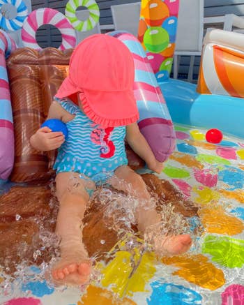 Seorang bayi yang mengenakan topi matahari dan baju renang bertema kuda laut duduk di kolam tiup berwarna-warni sambil memercikkan air