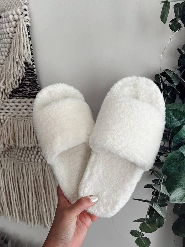 Person holding a pair of fluffy white slippers, suitable for home use, near a woven wall decoration and green foliage
