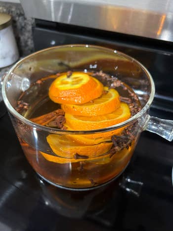 A wide   cookware  connected  a stove contains simmering h2o  with sliced oranges and cloves, suggesting a homemade simmering fragrance oregon  mulled beverage