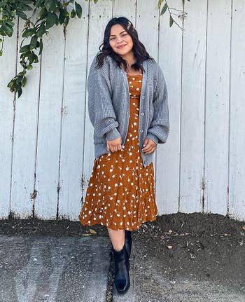 Person wearing a patterned dress, chunky knit cardigan, and boots, stands in front of a wooden fence with vines above. Casual fall fashion