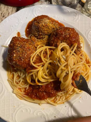 Spaghetti with tomato sauce and three large meatballs on a white plate, topped with grated cheese. Fork twirls pasta