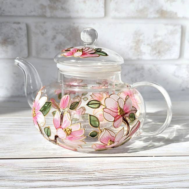 Glass teapot with floral plan featuring pinkish flowers and greenish leaves, placed connected a airy woody surface