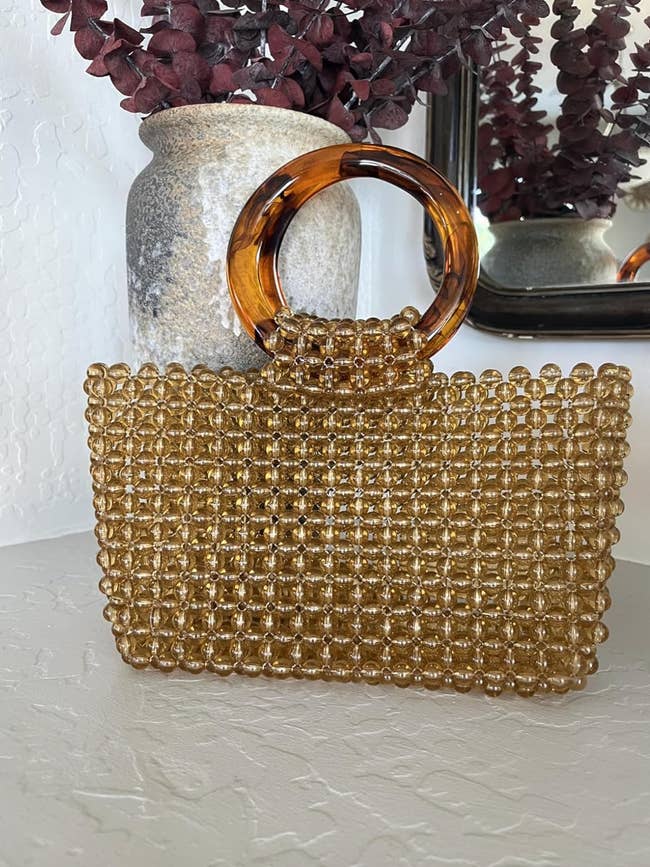 Woven handbag with a circular grip  connected  a shelf, adjacent  to a vase. Suitable for trendy, chic shopping