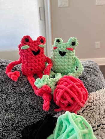 Two cute, handmade crochet frog toys sit on a blanket with yarn balls nearby. Perfect for gifting or as adorable home decor accents