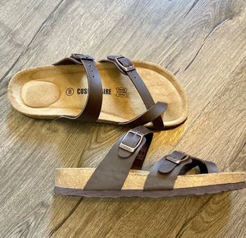 a reviewer's pair of cork sole sandals with adjustable straps on a wooden floor
