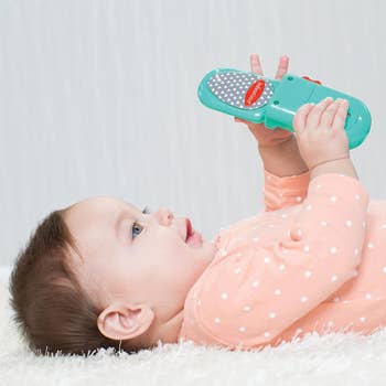 Baby lying down holding a toy flip phone, looking engaged and curious. The toy is colorful and features a large button and a textured screen