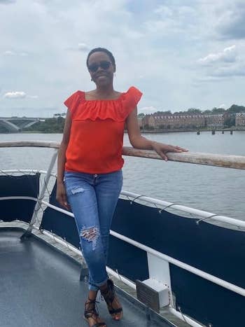 Person on a boat wearing a red off-shoulder top, ripped jeans, and sunglasses with a calm river and buildings in the background