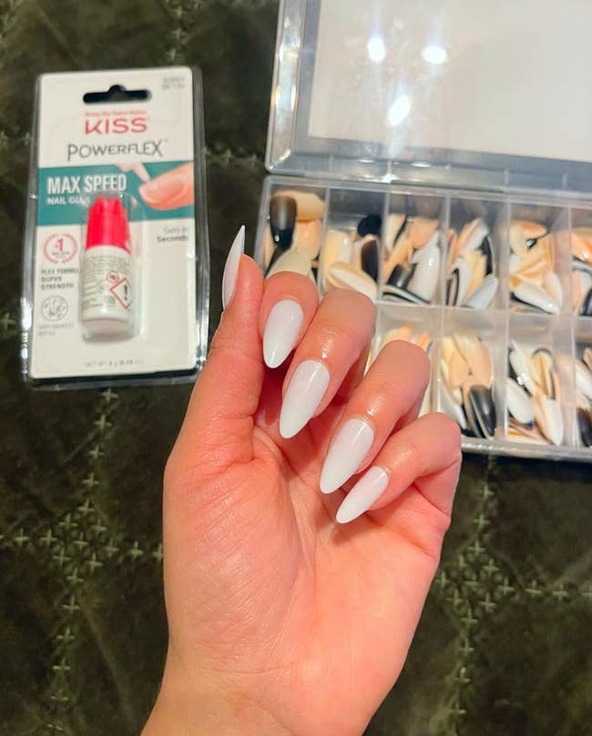 A manus  displaying long, white, coffin-shaped nails with a bundle  of KISS PowerFlex nail glue and a container  of assorted  fake nails successful  the background