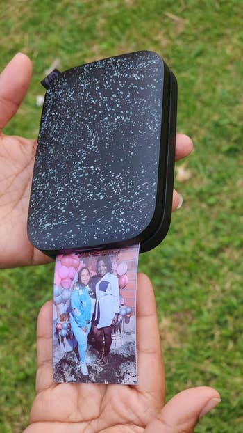 Portable photo printer held over grass, printing an image of two people standing in front of pink balloons