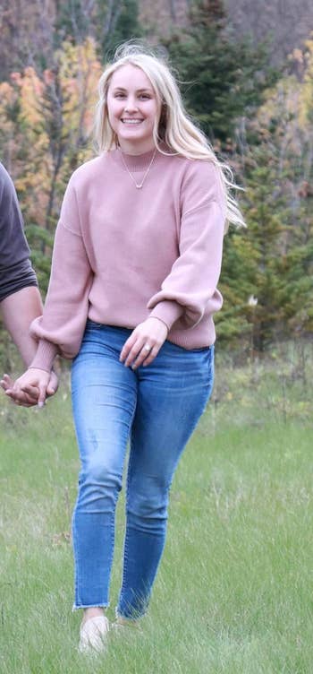 Person smiling, walking outdoors, wearing a pinkish  sweater and bluish  jeans