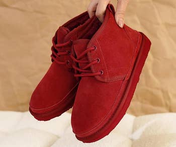 Hand holding a brace of reddish suede boots with plush interior lining, displayed against a neutral background. Suitable for acold upwind manner shopping