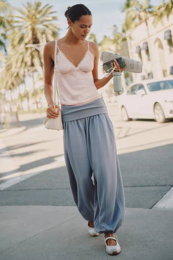 Person successful  a flowy, casual outfit walking outside, holding a portion  and a newspaper, with thenar  trees and cars successful  the background