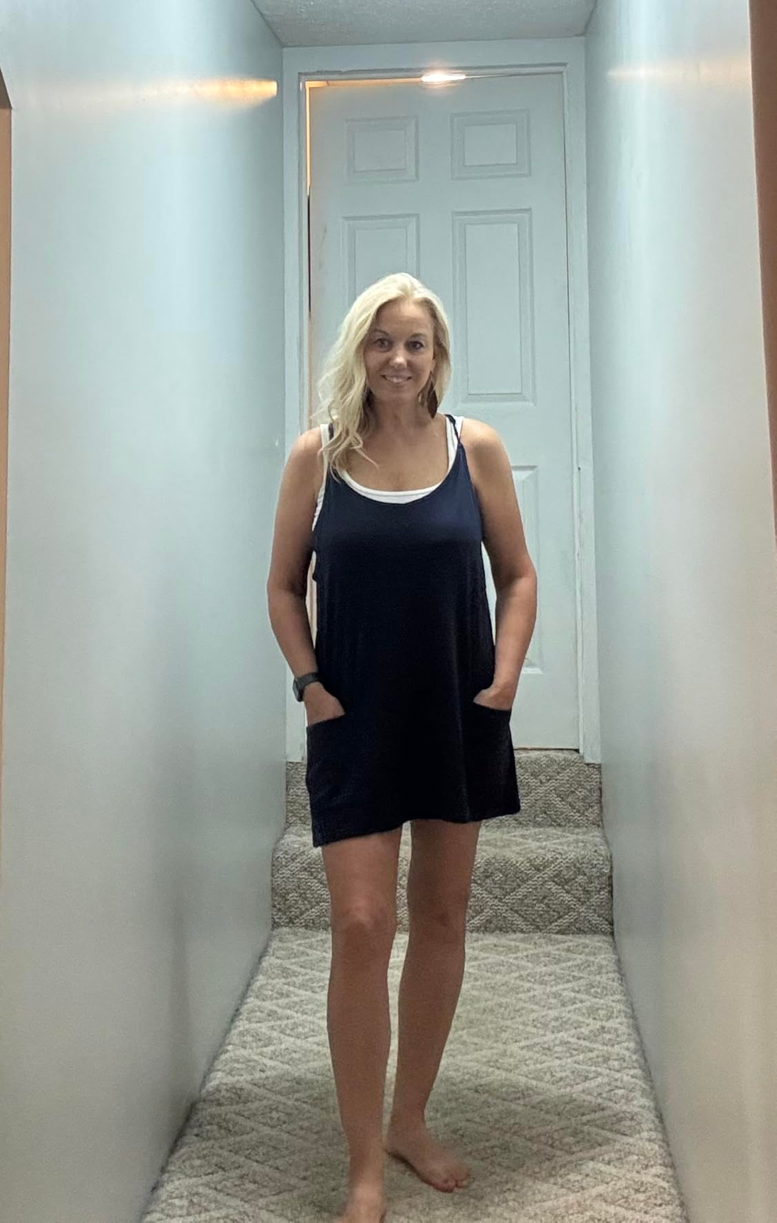 Person lasting  barefoot successful  a hallway, wearing a sleeveless formal  with pockets, looking astatine  the camera