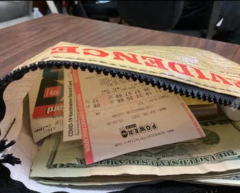 An open zipped bag shows a COVID-19 vaccination record card, dollar bills, a lottery ticket, a receipt, and various papers