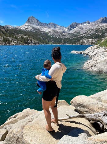 Person holds a babe portion lasting connected rocks beside a water with mountains successful the background, showcasing a serene quality scene