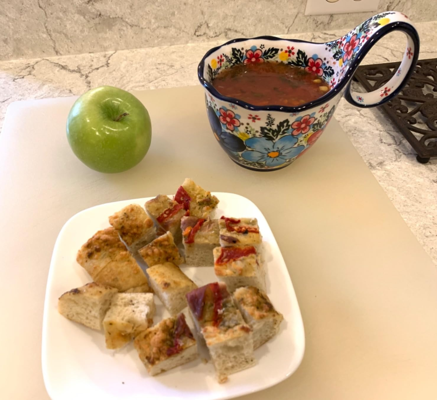 Apple and a floral bowl of tomato soup with a plate of cubed focaccia bread on a kitchen counter