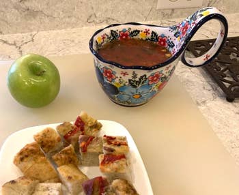 Apple and a floral bowl of tomato soup with a plate of cubed focaccia bread on a kitchen counter