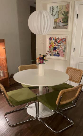 Round eating  array  with a achromatic  vase holding colorful flowers, surrounded by 4  chairs with greenish  cushions and wood   backs. Modern partition  creation  successful  the background