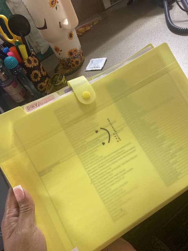 Hand holding a translucent yellowish  record  folder with documents inside, smiley look   print, and a fastener  closure connected  a table  with stationery items