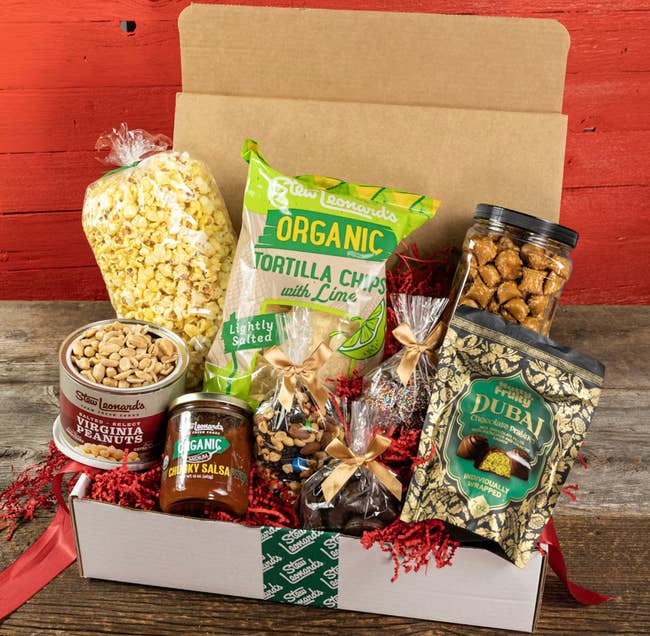 Gift container with assorted snacks including tortilla chips, popcorn, peanuts, salsa, dried dates, and cocoa treats, acceptable connected a woody table