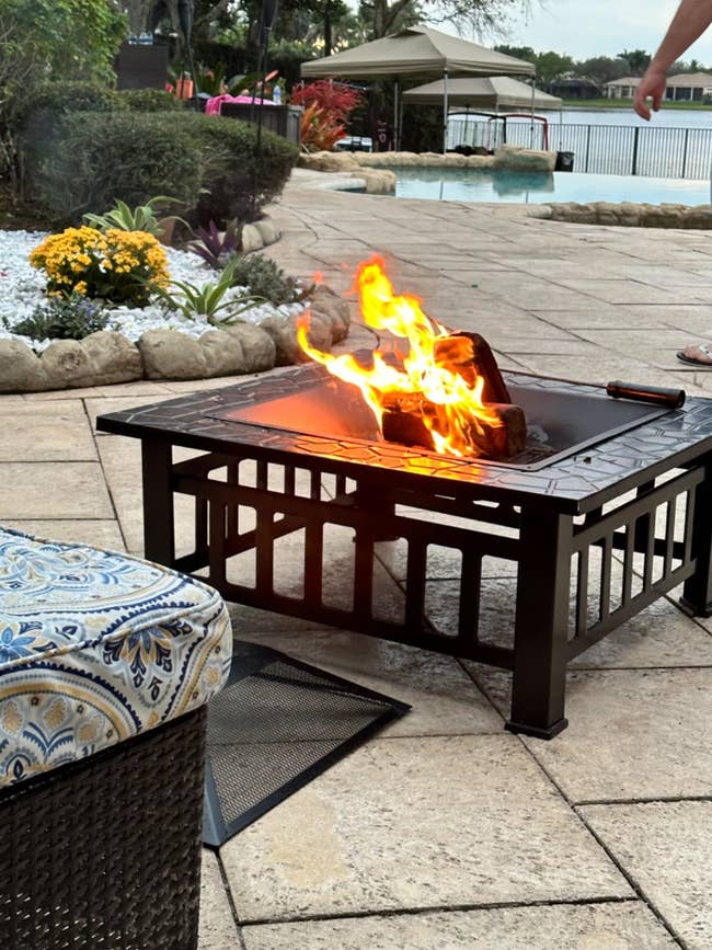 Outdoor fire pit on a patio near a pool, with patterned seating in the foreground. No persons are visible
