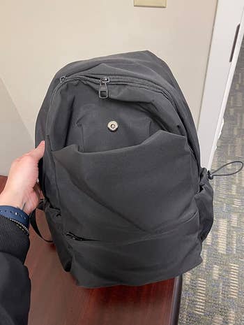 Person holding a black backpack with multiple compartments, showcased on a table in a shopping article. No other identifiable persons are in the image