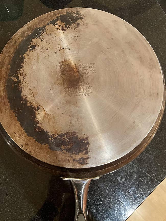 Well-used stainless steel frying pan on a stovetop, half of which is shiny and clean and the other half is stained