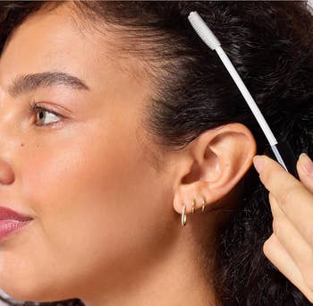 Person brushing their hairline with a mascara wand for styling, wearing tiny  hoop earrings