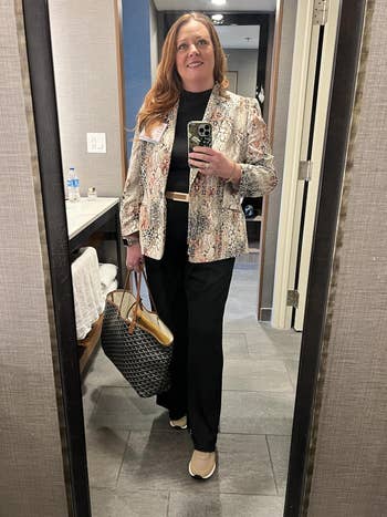 Person in mirror selfie wearing a patterned blazer, black outfit, and holding a large tote bag, standing in a bathroom